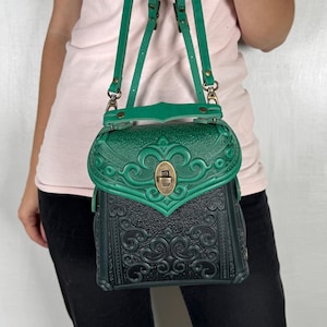 Könnte beinhalten: Ein grüner Lederrucksack mit dekorativer Klappe und Messingfarbenem Verschluss. Die Tasche hat geprägte Blumenmuster und verstellbare grüne Riemen. Die Tasche wird von einer Person gehalten.