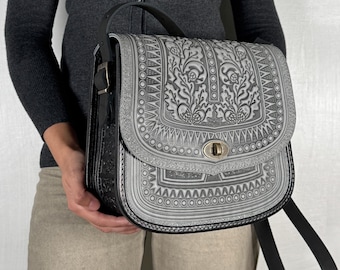 May include: A gray and black leather satchel with a decorative floral pattern. The bag has a flap closure with a metal clasp, a black shoulder strap, and a black side panel. The bag is held by a person wearing a gray cardigan and tan pants.