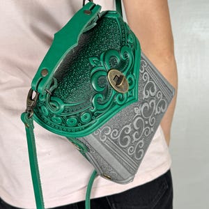 Könnte beinhalten: Eine grüne und graue Leder-Umhängetasche mit aufwendigen geprägten Designs. Die Tasche hat eine dekorative Klappe mit einem Verschluss und einen langen, verstellbaren Riemen. Das Design umfasst florale und scrollartige Muster.