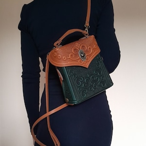 Op de afbeelding: Een groene en bruine leren tas met ingewikkelde bloemmotieven. De tas heeft een bovenklep met een decoratieve metalen sluiting en een lange verstelbare band.