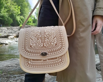 May include: A light brown leather shoulder bag with intricate floral embossed details. The bag has a flap closure and a long adjustable strap.