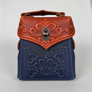 Puede incluir: Un pequeño bolso de mano de cuero azul y marrón con intrincados diseños florales en relieve. El bolso tiene un cierre de solapa superior con un broche de metal y una asa corta.