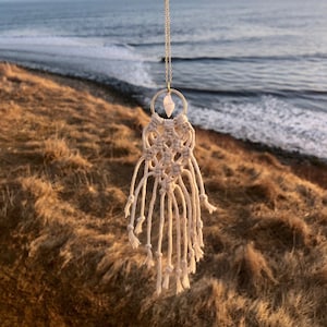 May include: A white macrame necklace with a crystal pendant hangs against a backdrop of a sandy beach and ocean waves.