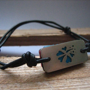 Op de afbeelding: Een zwart leren armband met een zilveren bedel met een blauwe hibiscusbloem met een uitsnijding. De bedel is met een knoop aan de armband bevestigd.