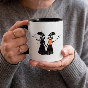 May include: A white ceramic mug with a black rim and handle, featuring a graphic illustration of two witches holding hands under umbrellas, surrounded by a swarm of black bats flying across the surface of the mug against a plain white background.