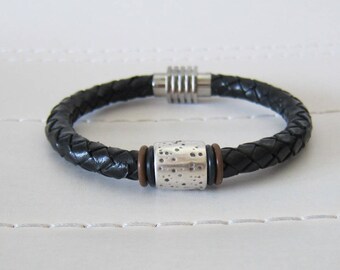 May include: A black leather bracelet with a braided design and a silver magnetic clasp. The bracelet features a silver bead with a textured surface.