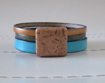 May include: A multi-layered leather bracelet with a copper magnetic clasp. The bracelet is made of three different colors of leather: turquoise, brown, and gray.