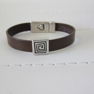 May include: A brown leather bracelet with a silver square charm featuring a spiral design.