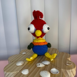 May include: A crocheted toy rooster with large eyes, red, yellow, orange, blue, and green colors. The rooster is sitting on a wooden flower-shaped stand with seashells.