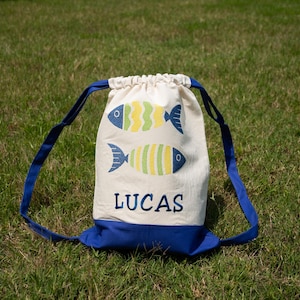 May include: A drawstring backpack with a cream-colored body and a blue base and straps. The bag features two embroidered fish in yellow, green, and blue, and the name "LUCAS" in blue.