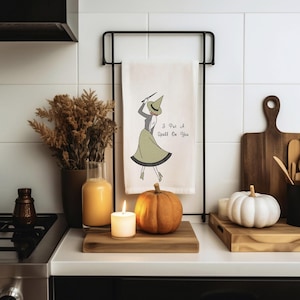 May include: A kitchen scene with a white towel featuring a witch illustration and the text "I Put A Spell On You." An orange pumpkin, white pumpkin, and lit candle sit on a wooden surface. Dried plants and a glass vase are also present.