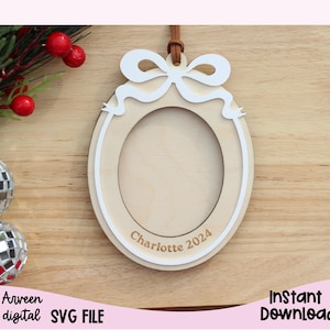 May include: A white wooden ornament shaped like a Christmas bauble with a bow on top. The ornament has a circular cutout in the centre for a photo and the text "Charlotte 2024" is written below the cutout.