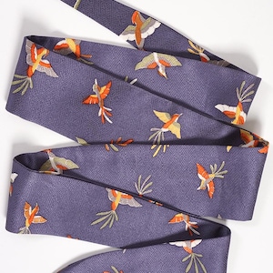 May include: A long, narrow purple scarf featuring a repeating embroidered pattern of small birds in shades of orange, yellow, and white. The fabric has a subtle, woven geometric texture throughout, and the scarf is folded into a layered shape.