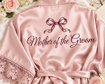 May include: A light pink satin robe featuring a dark red graphic of a bow and the text Mother of the Groom printed on the back. The robe has wide sleeves with intricate floral lace trim along the edges and a matching satin tie belt.