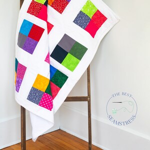 May include: A white quilt with colorful squares of fabric. The quilt is folded and draped over a wooden ladder.