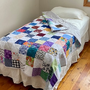 May include: A patchwork quilt featuring a range of colours and patterns, including floral, geometric, and abstract designs. The quilt is made from squares of different fabrics sewn together. The quilt is on a bed with a white sheet and a white pillow.
