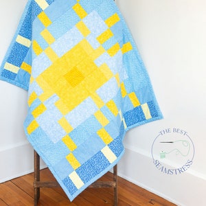 May include: A handmade quilt with a geometric pattern in shades of blue and yellow. The quilt features a central yellow square surrounded by blue and yellow rectangles. The quilt is draped over a wooden stand.