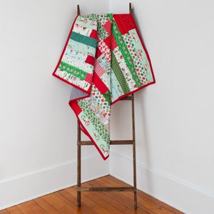 May include: A red and green patchwork quilt with a Christmas theme. The quilt is folded and draped over a wooden ladder.