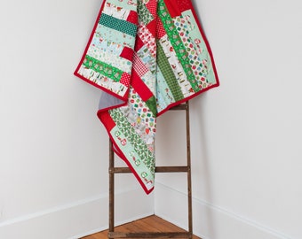 May include: A red and green patchwork quilt with a Christmas theme. The quilt is folded and draped over a wooden ladder.