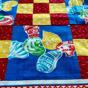 May include: A blue fabric with a patchwork design featuring red, yellow, and white squares. The center square has a design of seashells in various colors, including blue, green, yellow, and red.