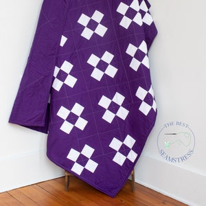 May include: Purple and white quilt with a geometric pattern of squares. The quilt is folded and draped over a small wooden stool. The text "THE BEST SEAMSTRESS" is visible in the lower right corner of the image.
