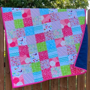 May include: A patchwork quilt with a pink, green, blue, and white floral pattern. The quilt is made of squares and has a pink border. The quilt is hanging on a wooden fence.