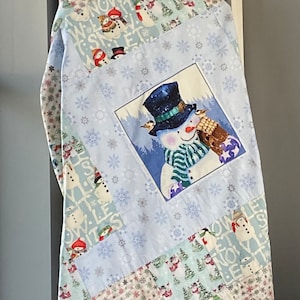 May include: A blue and white patchwork quilt with a snowman design. The snowman is holding a birdhouse and wearing a green and white striped scarf. The quilt is made of various fabrics with snowflakes and the words "Let It Snow" printed on them.