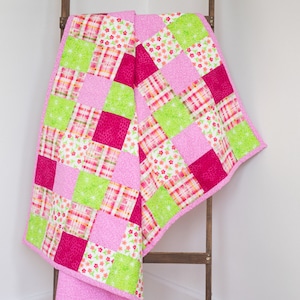 May include: A pink and green patchwork quilt with a variety of floral and geometric patterns. The quilt is folded and draped over a wooden ladder.