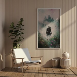 Puede incluir: Una impresi&oacute;n enmarcada de un paisaje de fantas&iacute;a con una figura vestida de negro caminando hacia un castillo en la distancia. La impresi&oacute;n se coloca contra una pared marr&oacute;n claro con una silla blanca y dos peque&ntilde;os jarrones blancos.