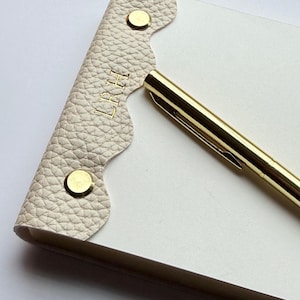 May include: A cream-colored notepad with a scalloped leather cover, secured with gold-colored rivets. The cover is embossed with the initials "LRH" in gold. A gold pen rests on the notepad.