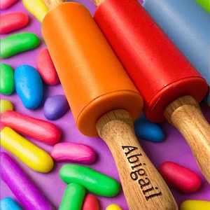 May include: Three wooden rolling pins with colorful silicone sleeves. The orange rolling pin has the name "Abigail" engraved on the handle. The rolling pins are surrounded by colorful play dough.