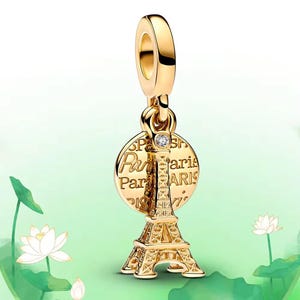 Könnte beinhalten: Goldfarbener Charm-Anhänger mit dem Eiffelturm und einem kreisförmigen Anhänger mit der Aufschrift "Paris". Der Charm enthält einen kleinen, klaren Edelstein und ist zum Anbringen an einem Armband oder einer Halskette bestimmt. Der Charm hängt an einer Schlaufe.