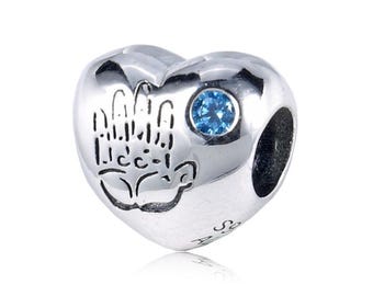 May include: A silver heart-shaped charm with a hand design and a small blue gemstone. The charm has a polished finish and a small hole for threading onto a bracelet or necklace. The charm is a jewelry accessory.