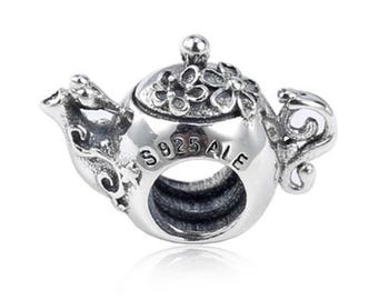 May include: A silver charm in the shape of a teapot. The charm has floral detailing on the lid and spout, with the text "S925 ALE" visible on the body. The charm is designed to be worn on a bracelet or necklace.