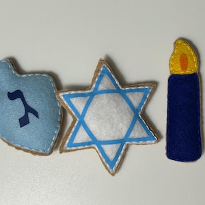 May include: Three felt decorations for Hanukkah: a blue dreidel with the Hebrew letter 'nun' on it, a white six-pointed star with a blue outline, and a blue candle with a yellow flame.