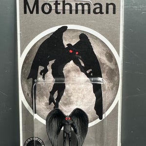 Könnte beinhalten: Eine Mothman-Figur in einer transparenten Kunststoffverpackung. Die Verpackung hat einen grauen Hintergrund mit dem Wort "Mothman" oben. Die Figur ist schwarz mit roten Augen und Flügeln. Die Verpackung trägt auch die Aufschrift "Winged Humanoid".