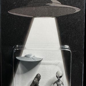 May include: A packaged toy set featuring a UFO, a Bigfoot figure, and a gray alien figure. The package has the word "BELIEVE" in green letters with a small alien icon. The UFO is emitting a bright white light.