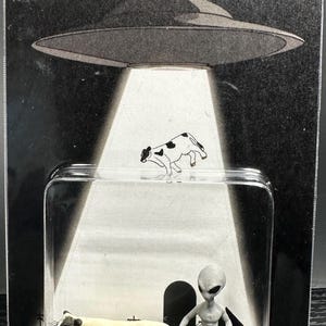May include: A packaged novelty item featuring a cow figurine, an alien figure, and the word "BELIEVE" in green. The background depicts a UFO and a spotlight effect, with a small cow graphic.