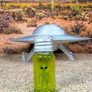 May include: A small clear glass bottle with a silver screw-top lid containing a bright neon green liquid and a miniature green alien figurine submerged inside. The bottle is positioned upright, showcasing the alien figure clearly through the glass.