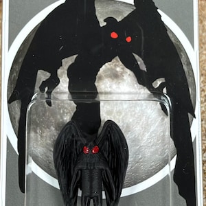 May include: A black Mothman figurine with red eyes stands inside a clear plastic blister pack. The backing card features a large moon graphic, the text Mothman at the top, and the word Humanoid at the bottom with a small circular logo.