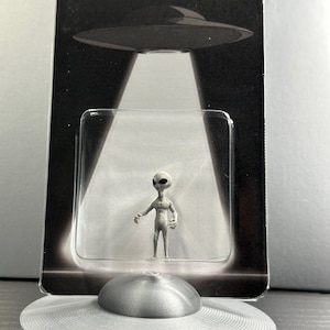 May include: A gray alien figurine stands in a clear plastic case, with a silver UFO model below. The packaging reads "BELIEVE" above a UFO graphic. The alien has a slender build and large eyes.