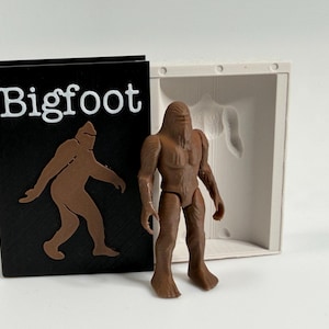Puede incluir: Una figura de acción marrón de Bigfoot está junto a una caja negra con la palabra "Bigfoot" en texto blanco y una silueta marrón de la criatura. Un contenedor blanco con un molde de Bigfoot está detrás de la figura.