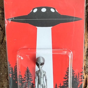 May include: A silver-coloured alien figurine encased in clear plastic packaging on a red backing card. The card features a black UFO, a white light beam, black pine tree silhouettes, and the text BELIEVE with an alien icon replacing the letter I.
