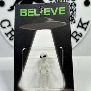 Puede incluir: Una pequeña figura de alienígena verde pálido dentro de un paquete de plástico transparente. El paquete tiene la palabra "BELIEVE" en letras verdes sobre una ilustración de un OVNI. El alienígena tiene grandes ojos negros y está de pie en un haz de luz.