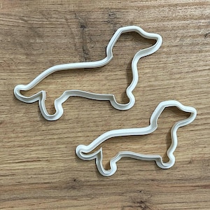 Puede incluir: Dos cortadores de galletas de plástico blanco con forma de perro salchicha. Los cortadores tienen la forma de un perro salchicha, con un cuerpo largo y patas cortas. Los cortadores están sobre una superficie de madera.