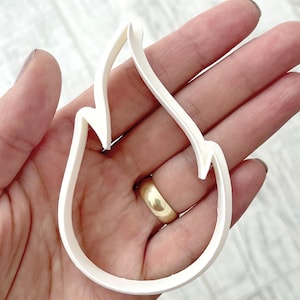 May include: A white plastic flame-shaped cookie cutter. The cutter is held in a hand, showing the shape and design. The cutter is a single piece, with a smooth, clean finish, ideal for baking.