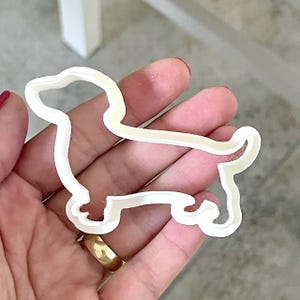 Puede incluir: Un cortador de galletas de plástico blanco con forma de perro salchicha. El cortador, sostenido en una mano, muestra el contorno del cuerpo, las patas y la cabeza del perro. Está diseñado para hornear y crear galletas con forma de perro.