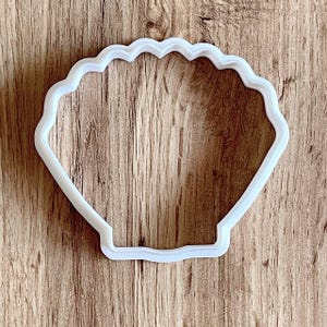 Puede incluir: Un cortador de galletas de plástico blanco con forma de concha. El cortador tiene un borde festoneado y un diseño sencillo. El cortador está colocado sobre una superficie de madera.