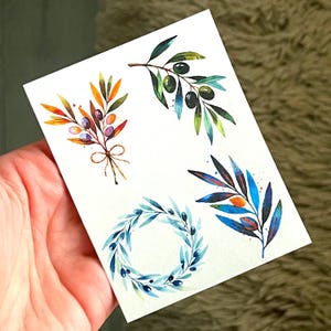 Peut inclure: Illustrations à l'aquarelle de branches et de couronnes d'oliviers. L'œuvre présente des olives et des feuilles vertes, bleues, oranges et violettes. Les dessins sont sur fond blanc.