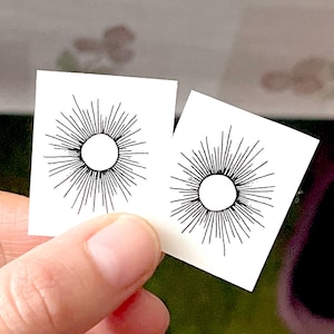 Op de afbeelding: Twee witte tijdelijke tatoeages met zwarte zonontwerpen. Elke zon heeft een cirkel in het midden en stralende lijnen.